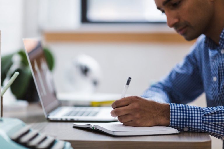 man writing on paper next to laptop