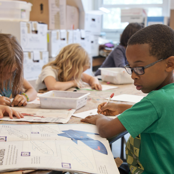 Captured in a metropolitan Atlanta, Georgia primary school, seated amongst his classmates, this photograph depicts a young African-American schoolboy who was in the process of drawing with a pencil on a piece of white paper. Note that the student was focused on a drawing book that referenced fantasy flying planes, while intent on creating his artwork, seemingly oblivious to all the classroom goings-on that surrounded him.