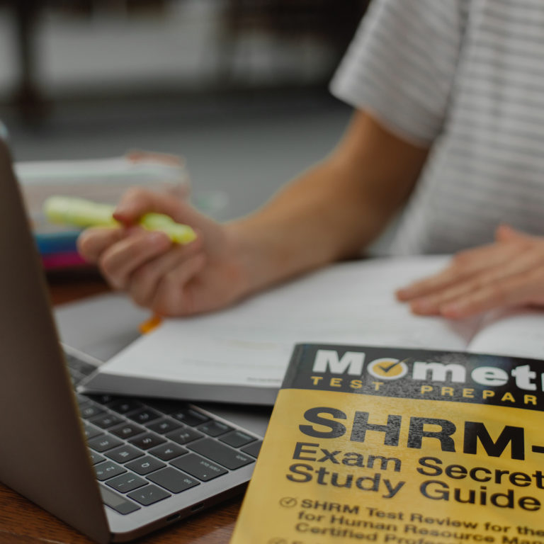 hand of a student behind a laptop highlighting text in a textbook with a yellow and black book in front entitled exam secrets study guide